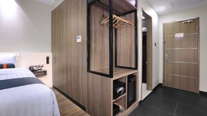 Een hotelkamer met een bed, een houten garderobe, een kledingroede, een klein plankje met een telefoon en een deur met een bordje.