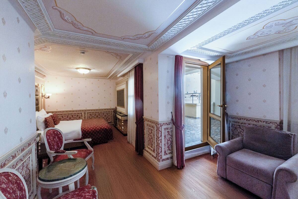 A room with a wooden floor, a sofa, a chair, and a bed with patterned wallpaper.