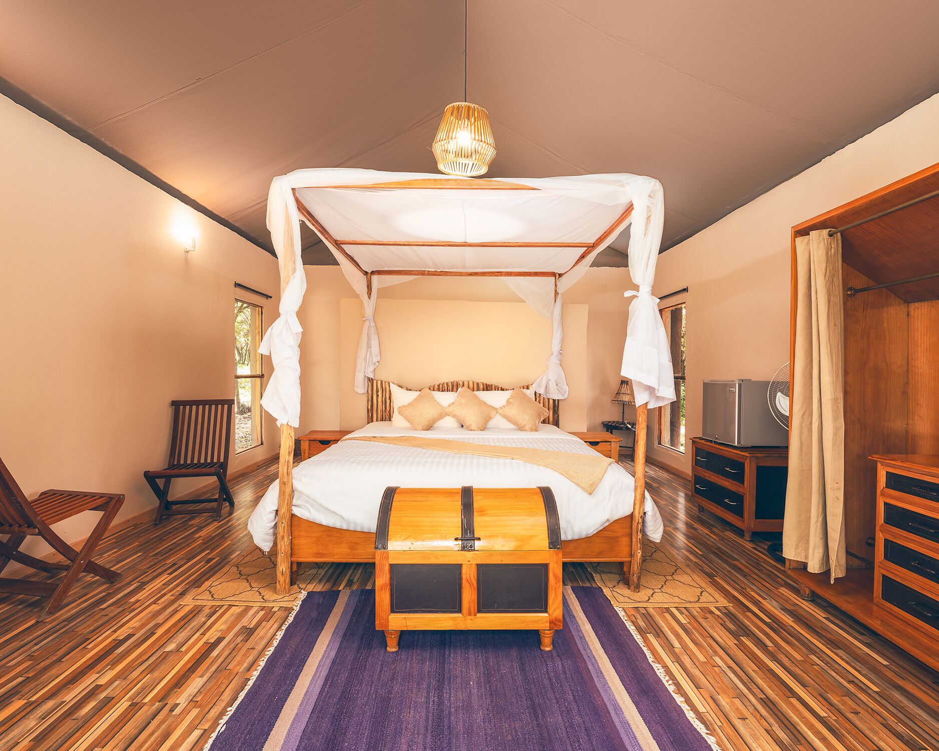 A four-poster canopy bed with a wooden frame and white bedding, a wooden bench at the foot of the bed, a small table beside the bed, and a television on a wooden cabinet.