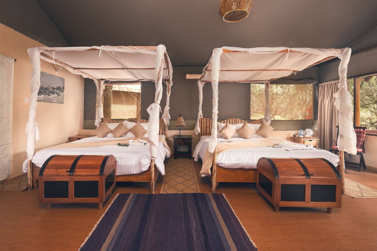 Een kamer met twee bedden onder hemelbedden, houten hoofdborden, een geweven hanglamp en uitzicht op bomen door het raam.
