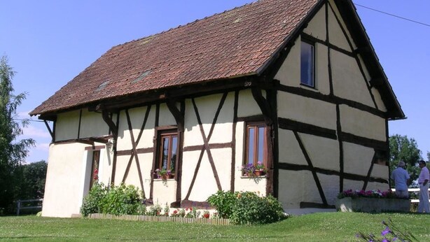 Una casa tradicional de entramado de madera con techo empinado, dos ventanas visibles con contraventanas y un pequeño jardín con flores y arbustos.