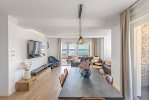 Uma sala de estar moderna com área de jantar, uma televisão de tela plana grande, um conjunto de sofás e vista para o mar.