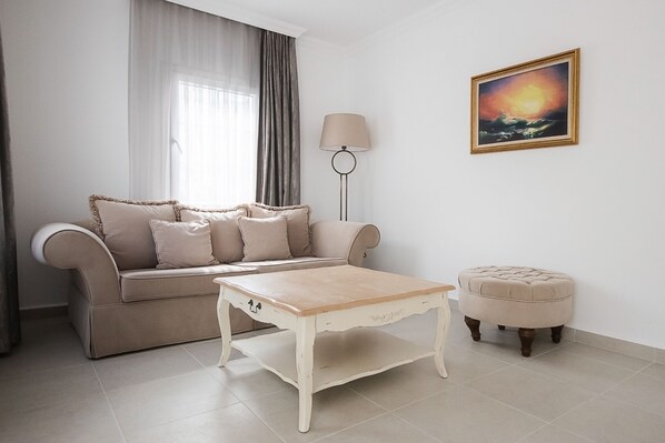 Una sala de estar con un sofá beige, una mesita de centro blanca pequeña, una lámpara de pie redonda y un cuadro enmarcado en la pared.