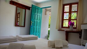 A bedroom with a bed, towels, a mirror, a teal door, and a window with a view of greenery. - Myndos Bed & Breakfast (Bodrum)