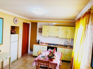 Una cocina compacta con armarios amarillos, una pequeña mesa de comedor con mantel a cuadros, un refrigerador y un reloj en la pared.