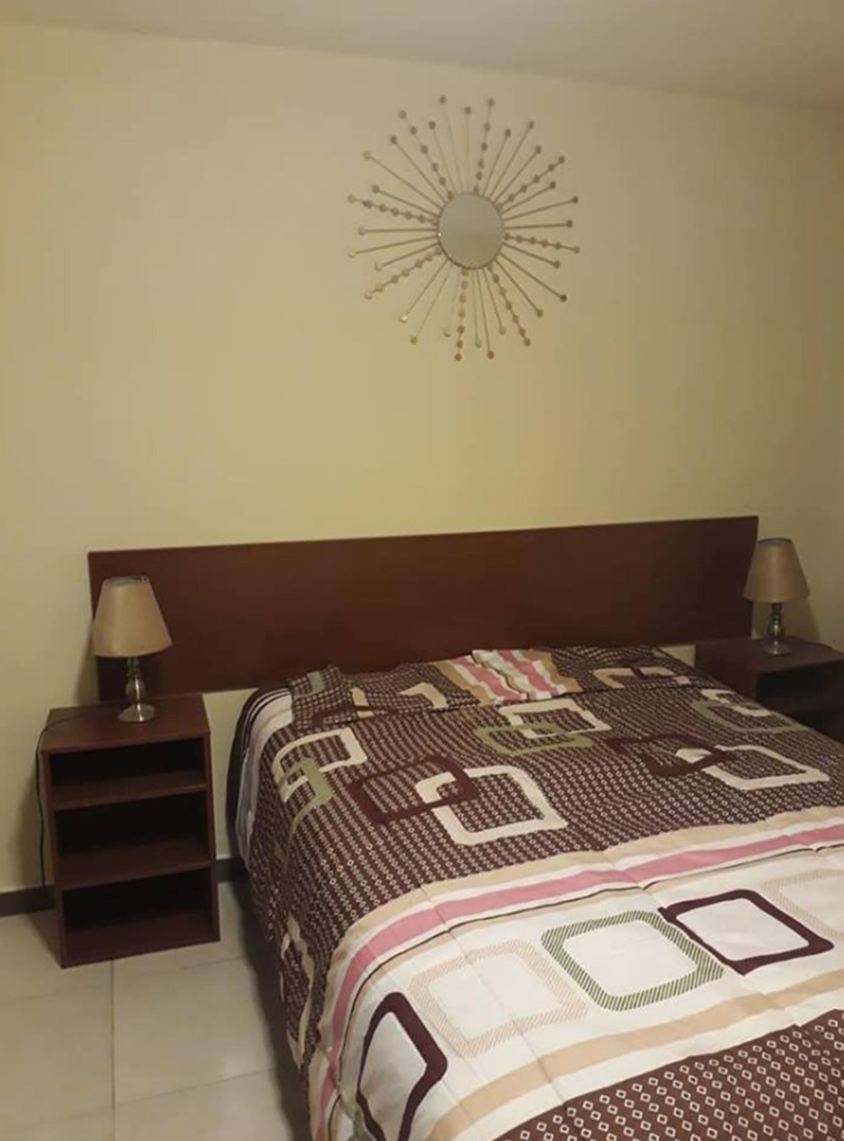 Un dormitorio con una cama, dos mesitas de noche con lámparas, un reloj de pared y una colcha estampada.
