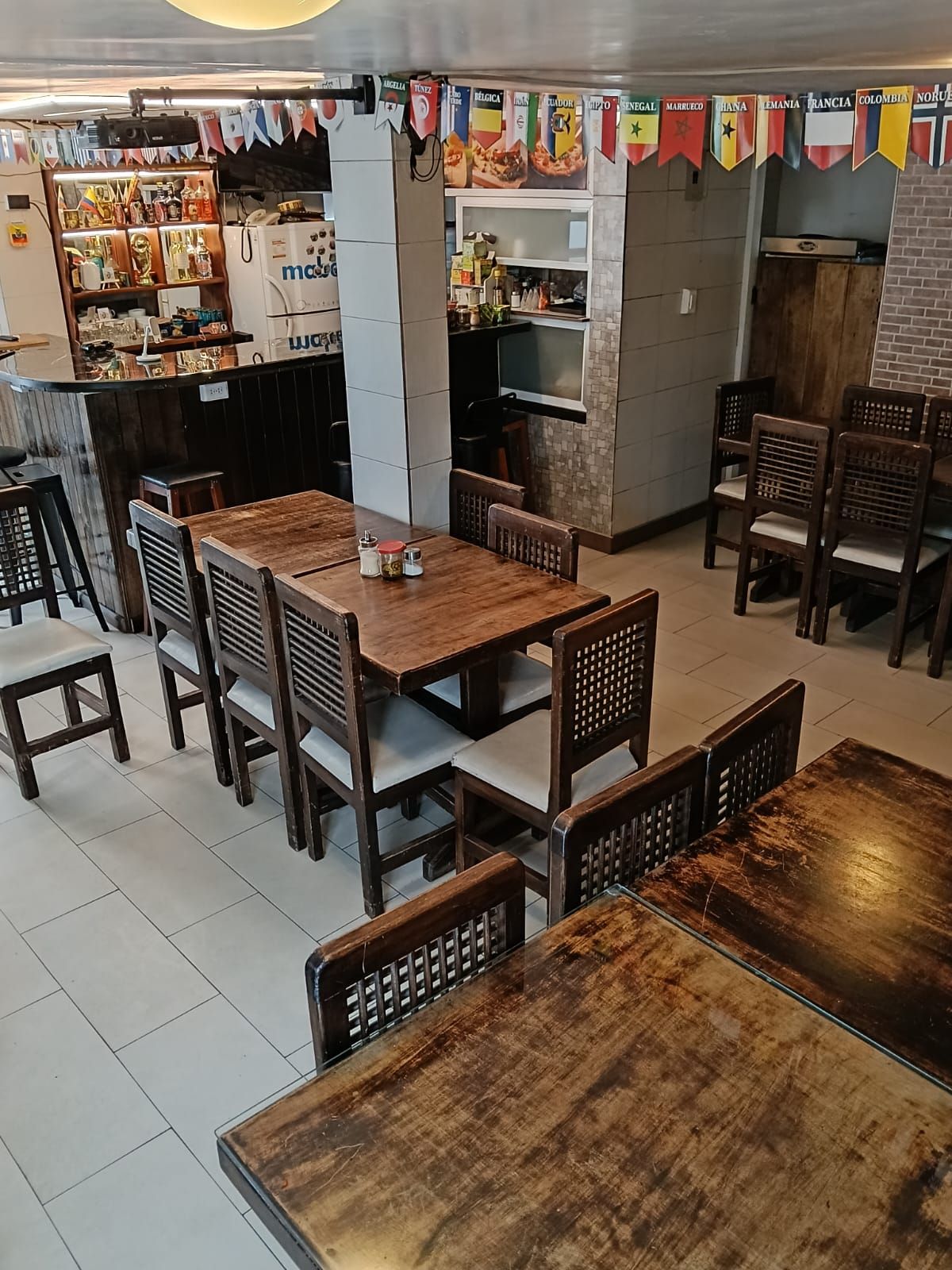 Un restaurante con mesas y sillas de madera, una zona de cocina y varias banderas colgando sobre la entrada.