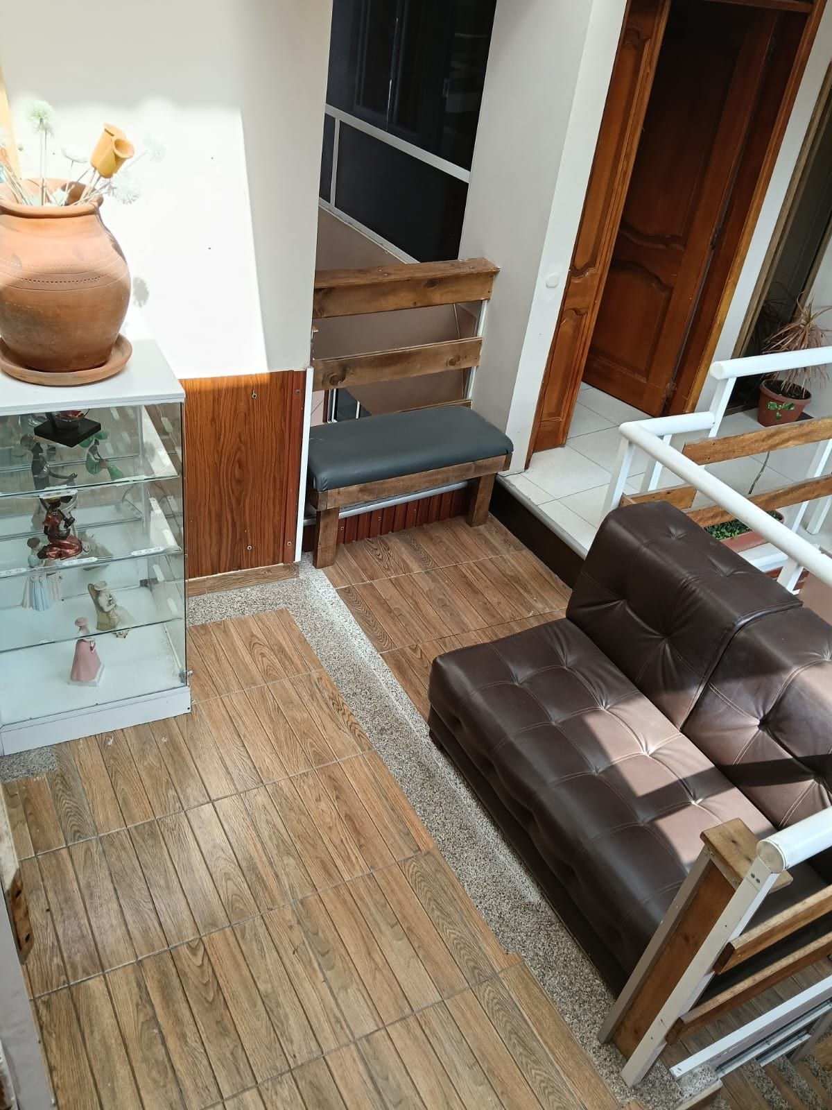 Een woonkamer met een bruine leren bank, een glazen vitrinekast, een houten bankje en een trap.