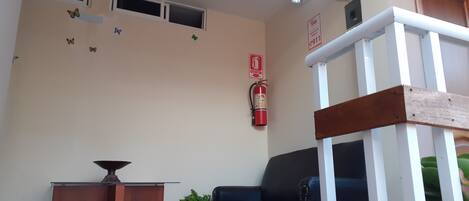 Un área de recepción con un escritorio de madera, un sofá de cuero negro, una planta en maceta, un extintor y una silla de madera.