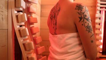 Eine Person sitzt auf einer Holzbank in einem Saunaraum mit einer Holzwand und weiteren Bänken.