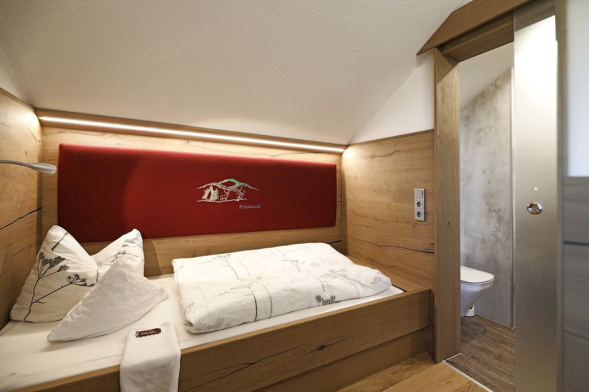 A small, compact room with a bed, a red headboard, a wooden bench, and a toilet.