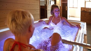 A person in a bathtub with visible tattoos, another person with a red ribbon, and a wooden interior. - Schätzlhof (Ruderting)