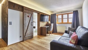A modern living room with a wooden floor, a black leather sofa, a wooden desk, and a large window with curtains. - Schätzlhof (Ruderting)
