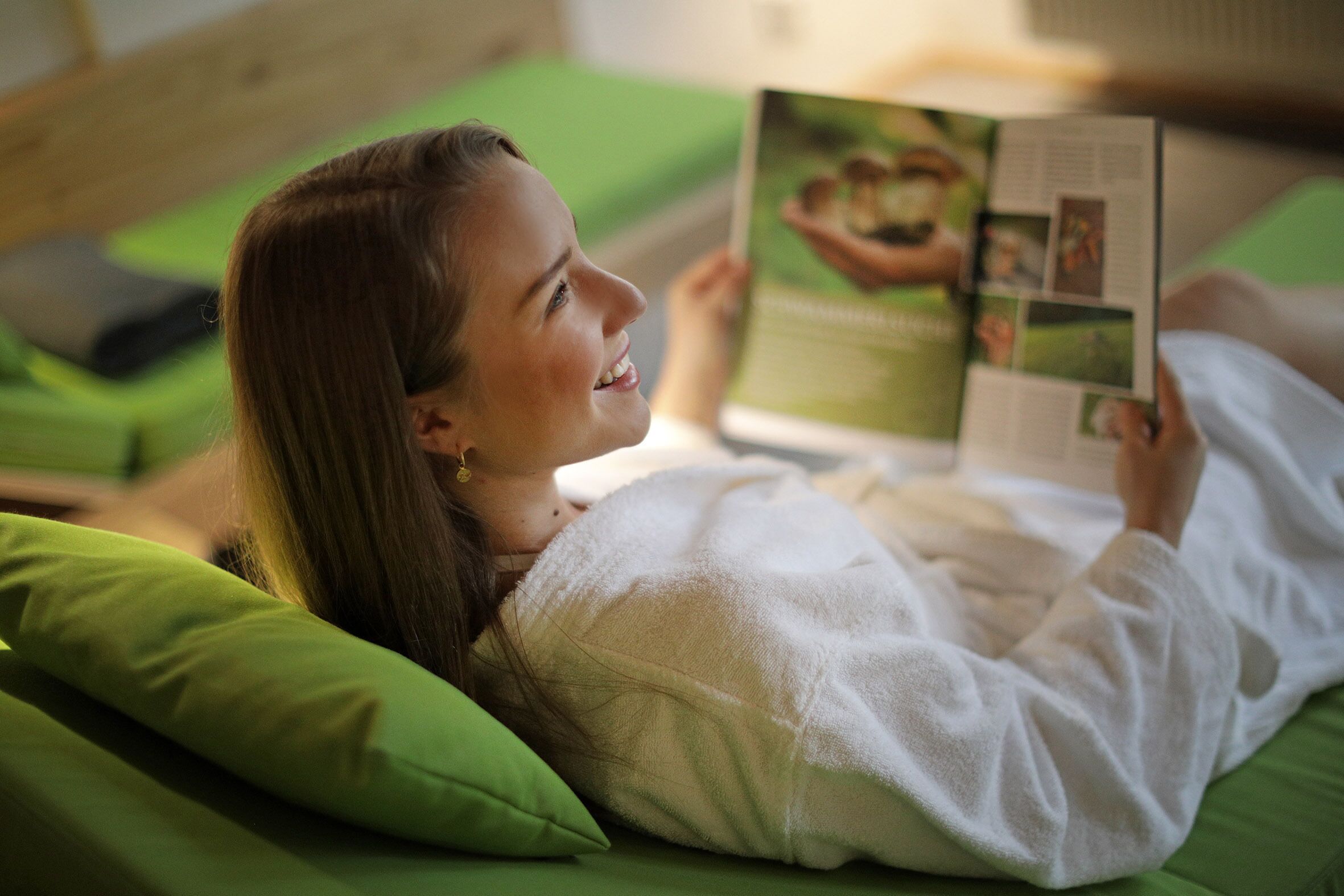 A person lying on a bed with green pillows, reading a magazine.