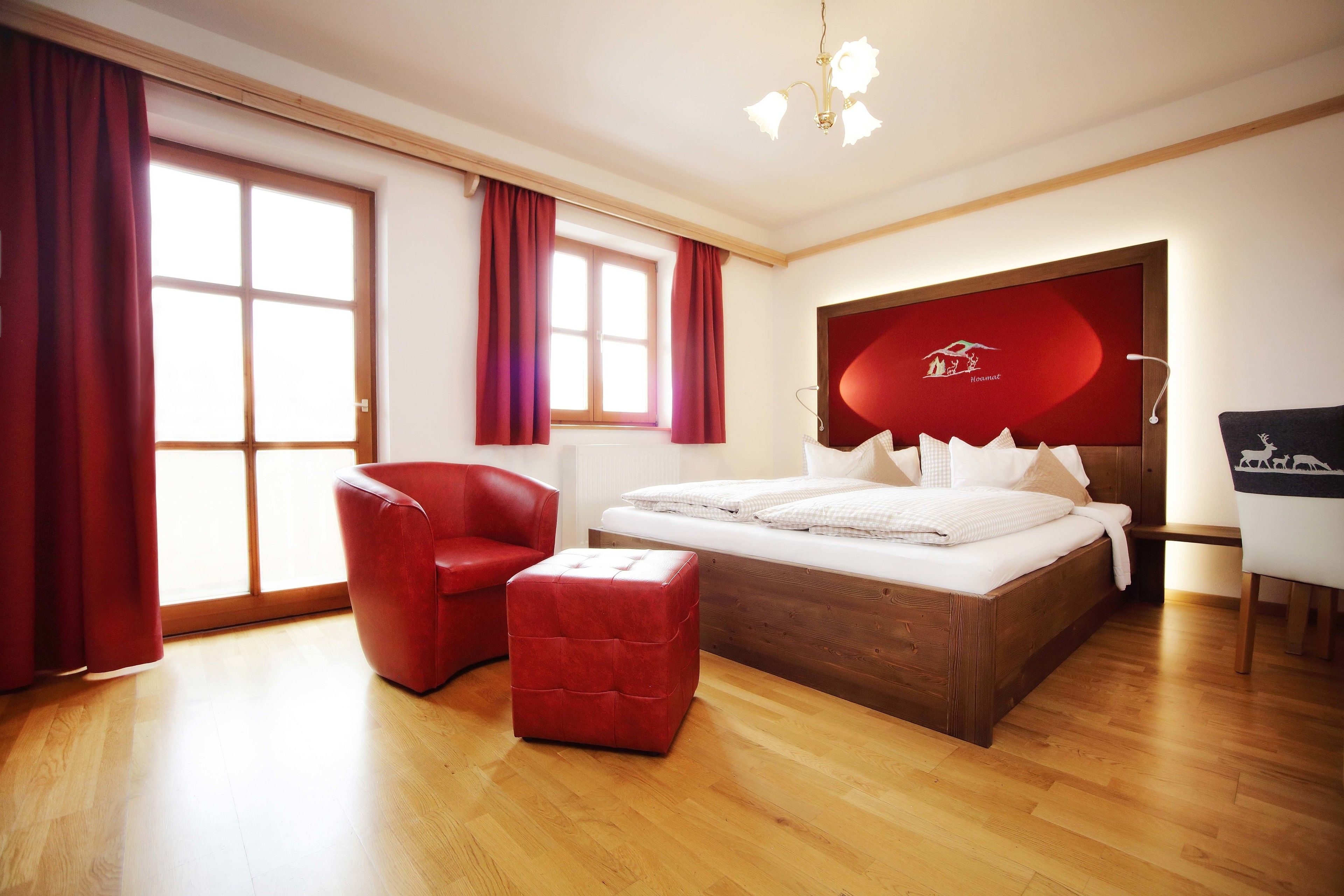 Quarto moderno com piso de madeira, uma cadeira estofada vermelha, um banquinho vermelho, uma cama com roupa de cama branca, uma cabeceira vermelha e uma escrivaninha com cadeira.