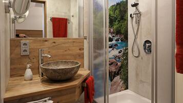 A modern bathroom with a wooden vanity, a large stone sink, a glass-enclosed shower, and a view of a rocky river.