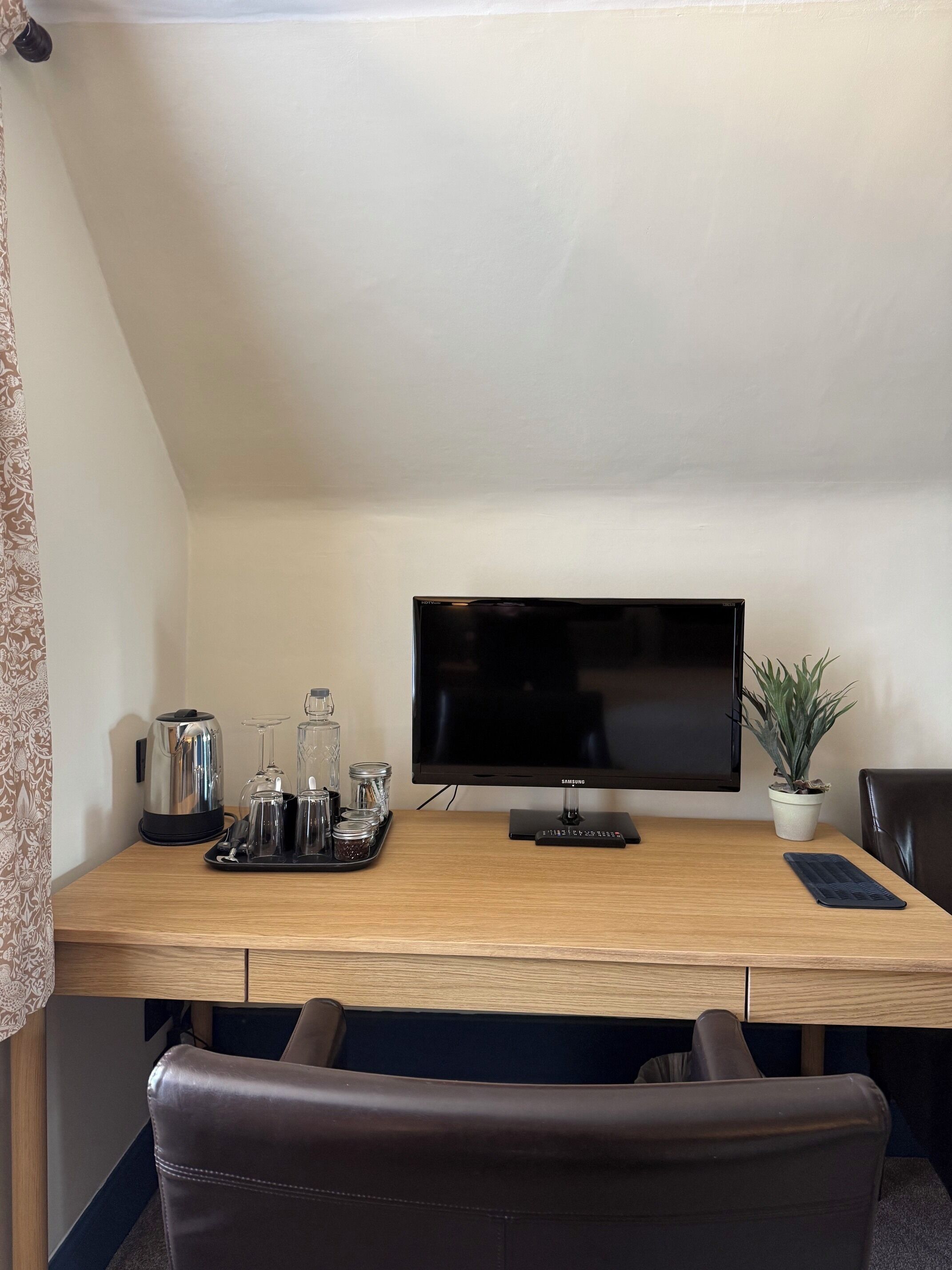 A hotel room with a wooden desk, a flat-screen TV, a potted plant, a leather chair, and a tray with a kettle, glasses, and jars.