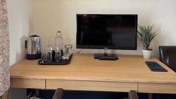 A hotel room with a wooden desk, a flat-screen TV, a potted plant, a leather chair, and a tray with a kettle, glasses, and jars.