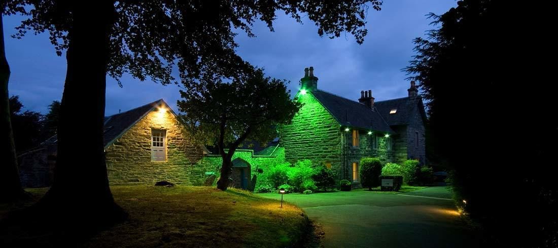 A stone building with a green-lit roof, surrounded by trees and a well-maintained lawn.