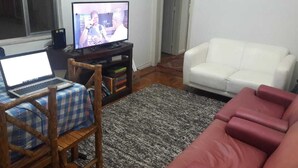Un salon avec un canapé blanc, un divan rouge, une chaise en bois recouverte d’une nappe à carreaux, un ordinateur portable posé sur une table et un téléviseur sur un meuble.