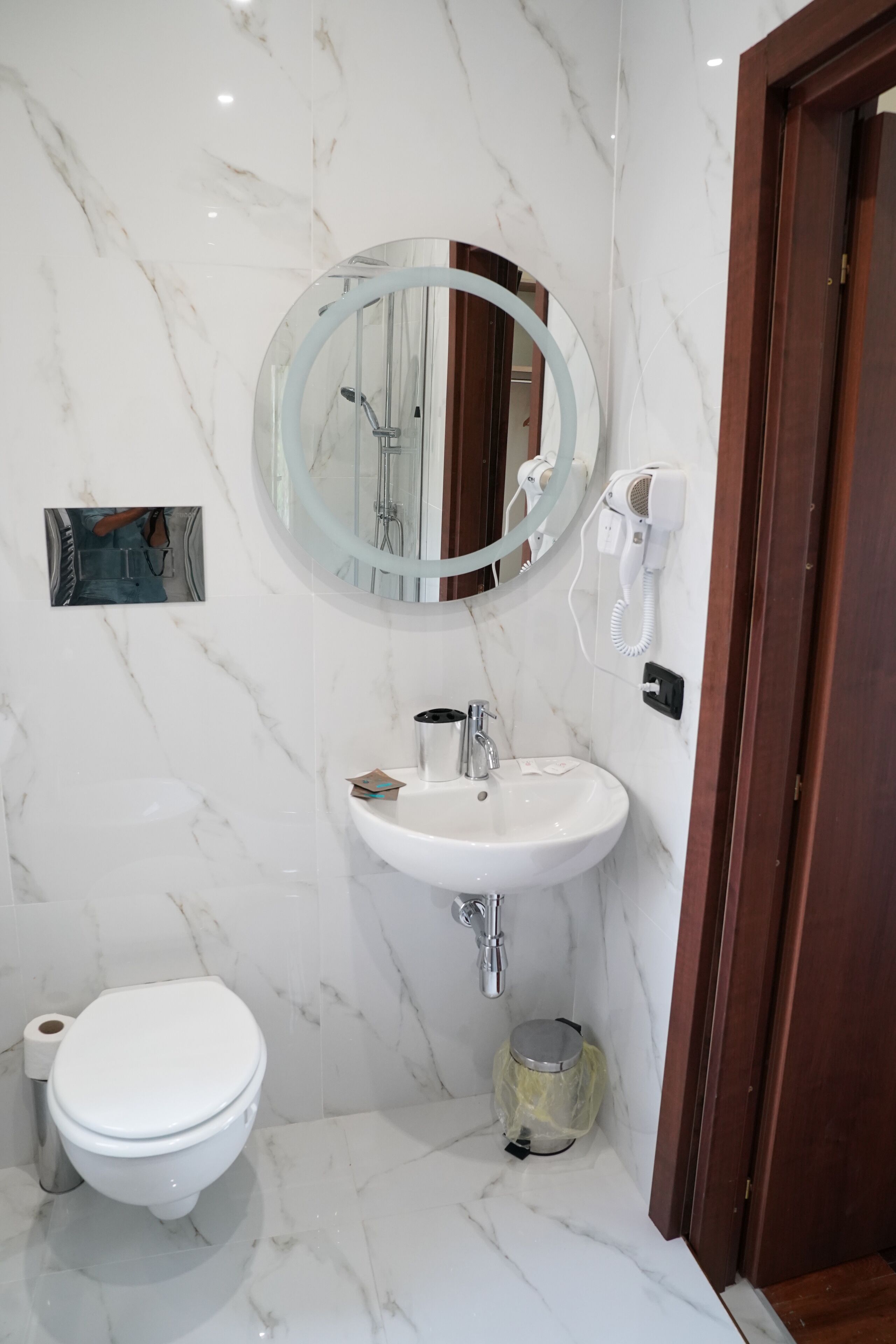 A bathroom with a marble floor, a white sink, a toilet, and a mirror with a light ring.
