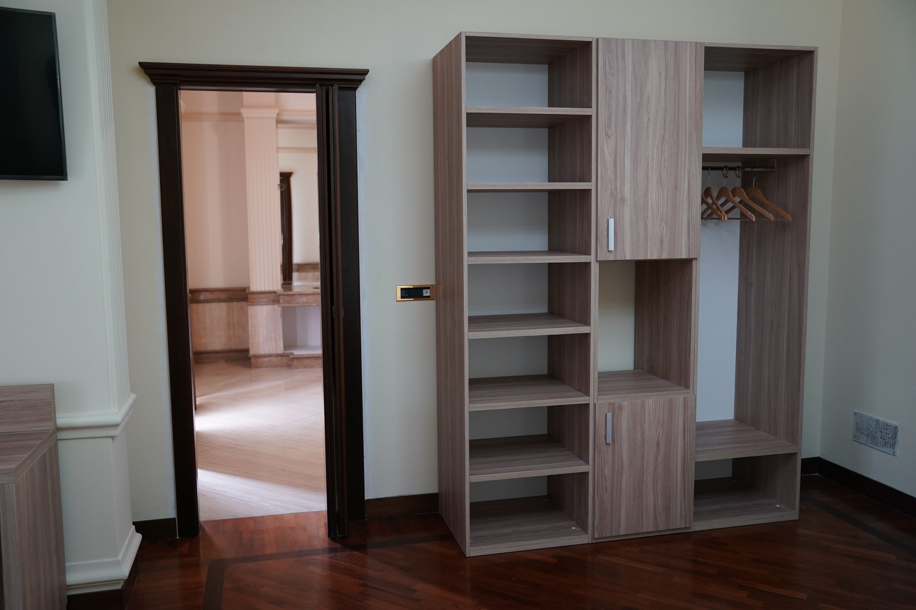 Una habitación con piso de madera, un armario con varios estantes y una puerta que da a otra habitación.