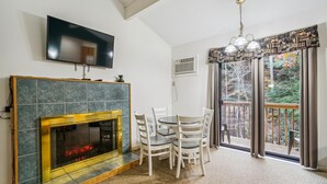 Una sala de estar con chimenea, una mesa de comedor con sillas, un televisor montado en la pared y un balcón con vistas a árboles.