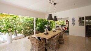 Een ruime eethoek met een houten tafel en rieten stoelen, omgeven door weelderige begroeiing en uitzicht op een zwembad.