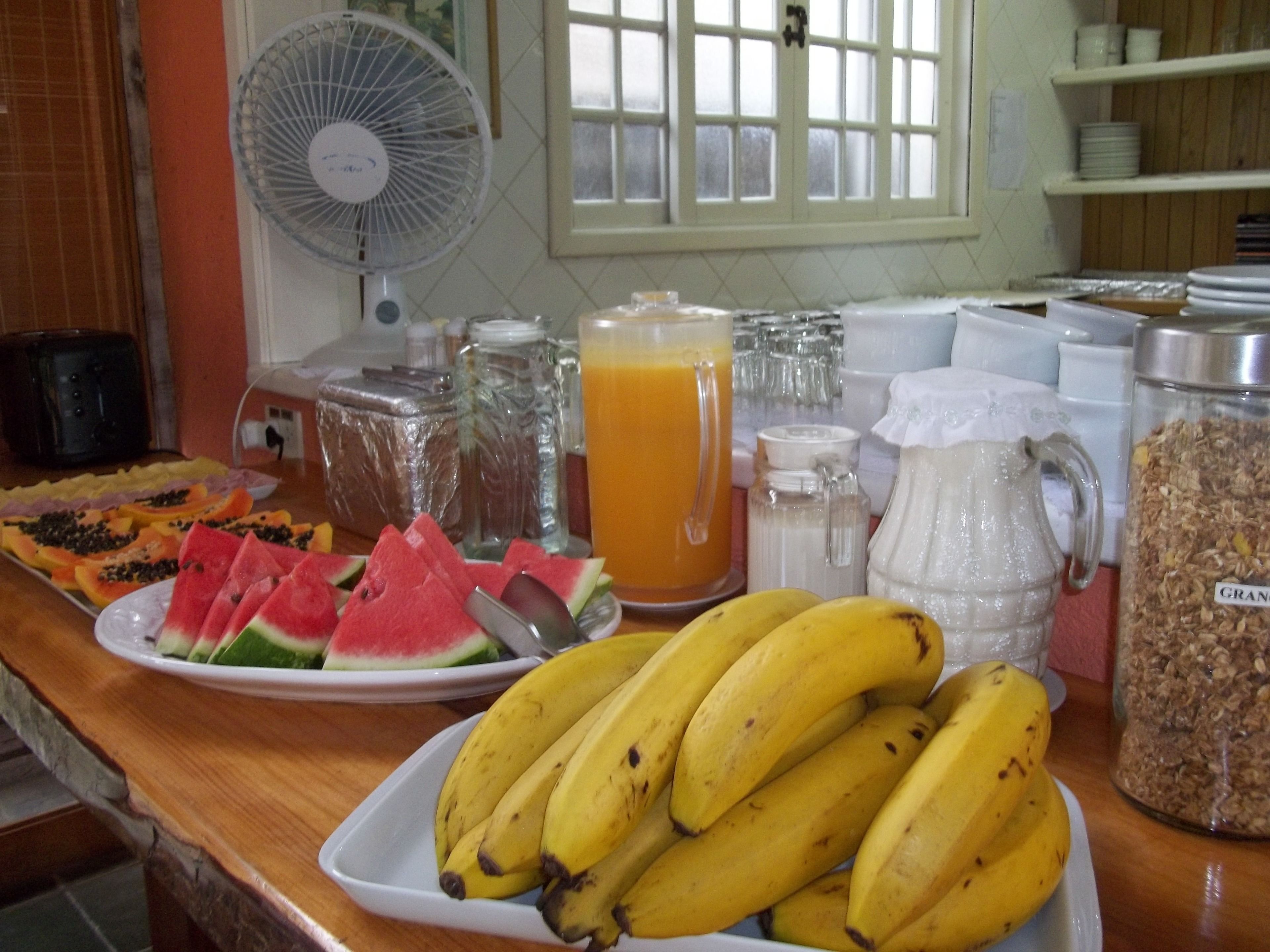 A breakfast buffet with various fruits, a pitcher of orange juice, and a fan in the background.