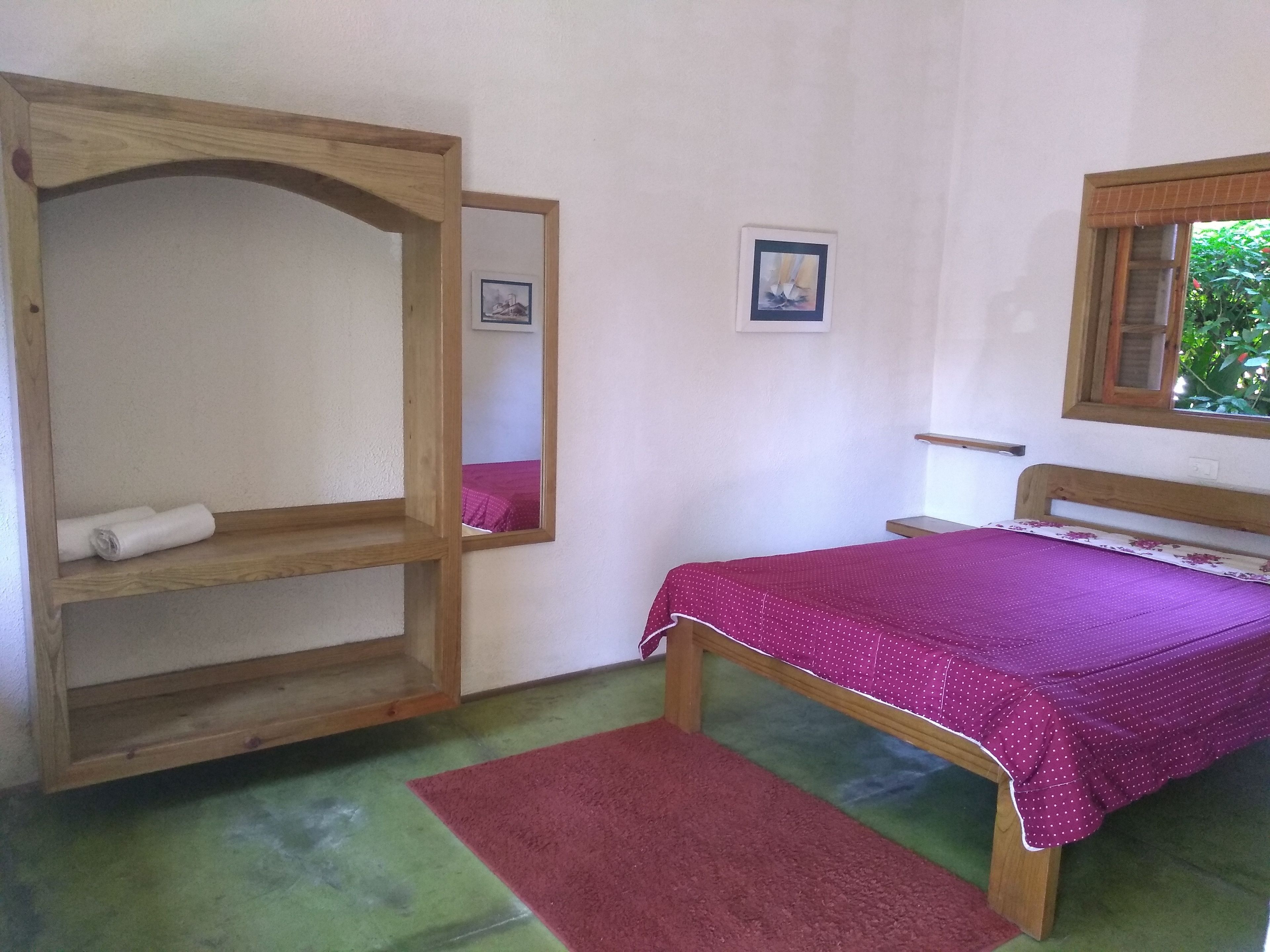 A bedroom with a wooden bunk bed, a single bed with a purple bedspread, a small bedside table, and a window with a view of greenery.