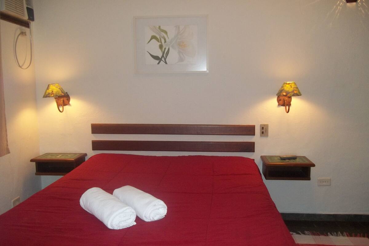 Una cama bien hecha con una colcha roja y dos toallas enrolladas, flanqueada por dos mesitas de noche con lámparas, y una obra de arte floral enmarcada en la pared.