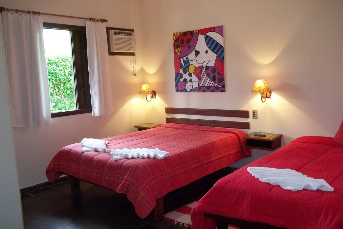 Habitación de hotel con dos camas, una ventana con cortinas, aire acondicionado de pared, un cuadro y dos lámparas de pared.