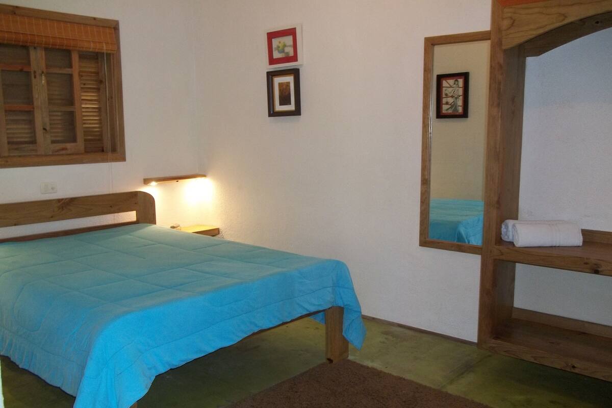 Una habitación con una cama, cabecera de madera, una mesita de noche con lámpara, un espejo y un estante con una toalla.