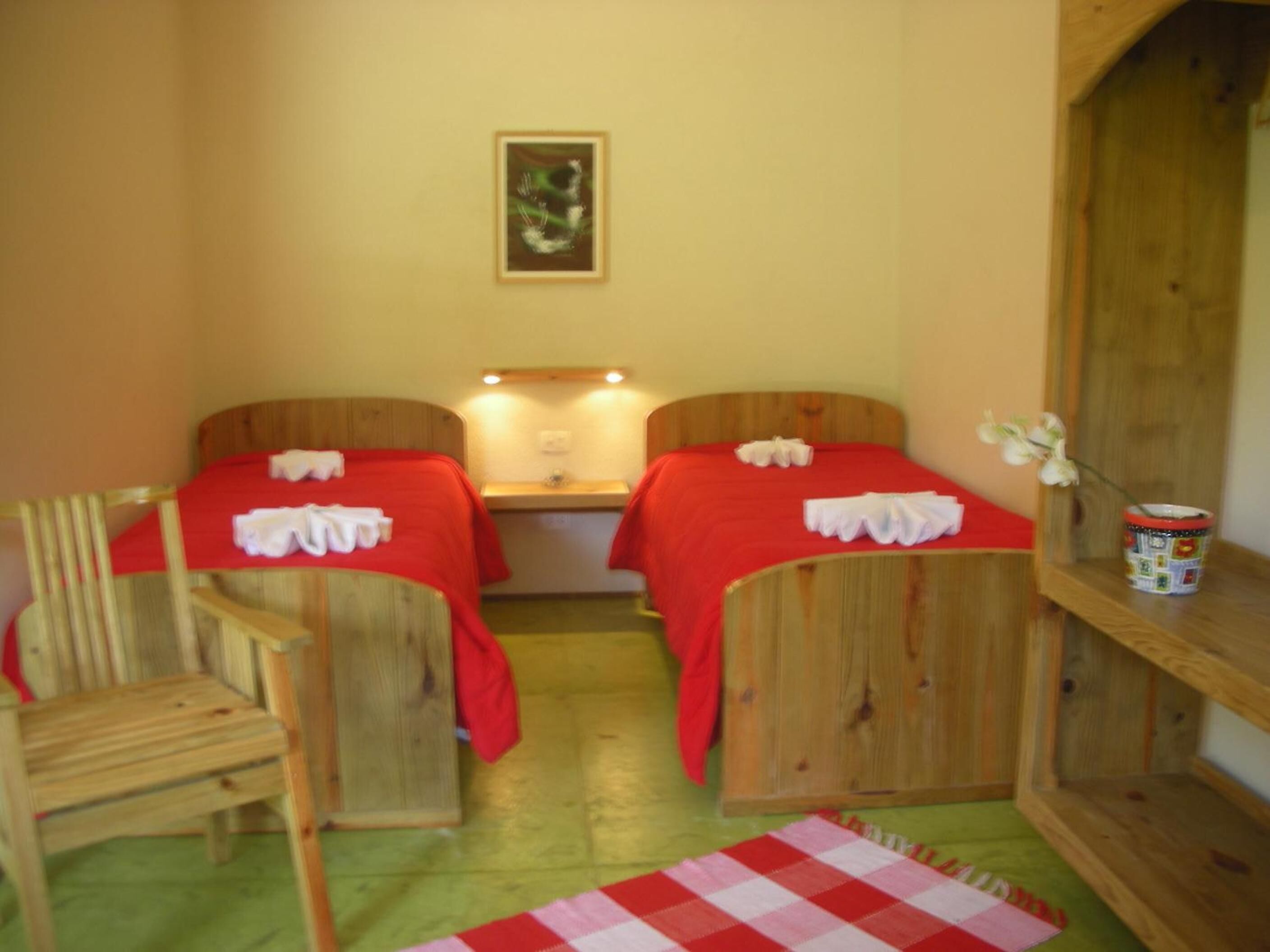 Una habitación con dos camas de madera, cada una con colcha roja y toallas blancas. Hay una silla de madera, una mesita con una lámpara, y una estantería con un objeto decorativo.