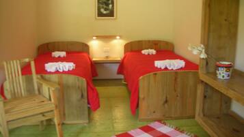 Una habitación con dos camas de madera, cada una con colcha roja y toallas blancas. Hay una silla de madera, una mesita con una lámpara, y una estantería con un objeto decorativo.