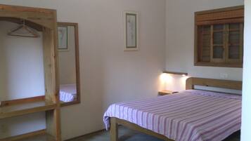 A bedroom with a single bed, a wooden wardrobe, a bedside table with a lamp, and a window with shutters.