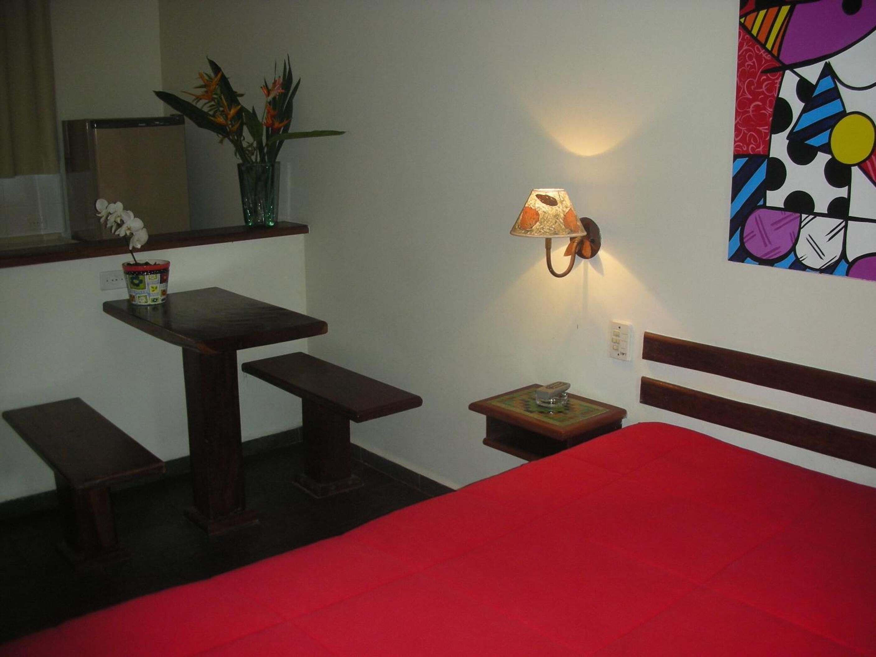 A small room with a bed, a table, and a bench. There is a lamp on the wall and a plant on the table.