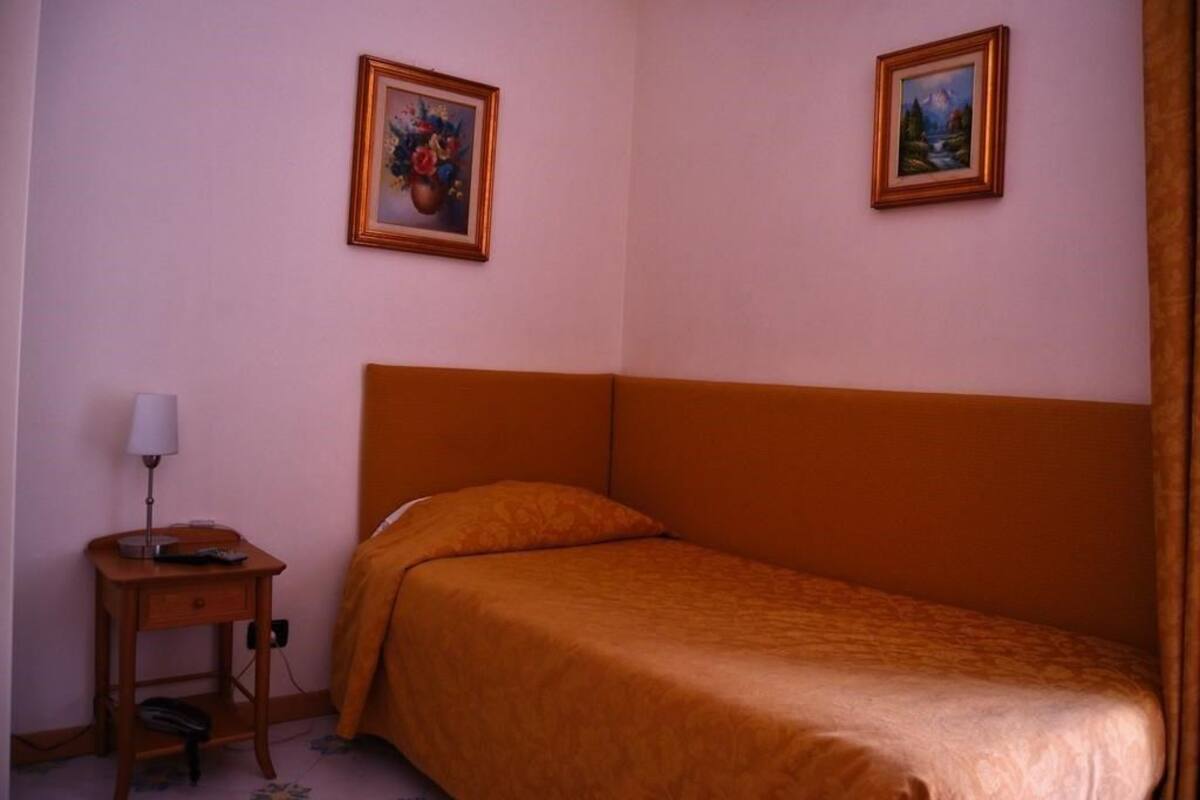 Quarto com uma cama de solteiro, uma mesa de cabeceira com abajur e duas quadros na parede.