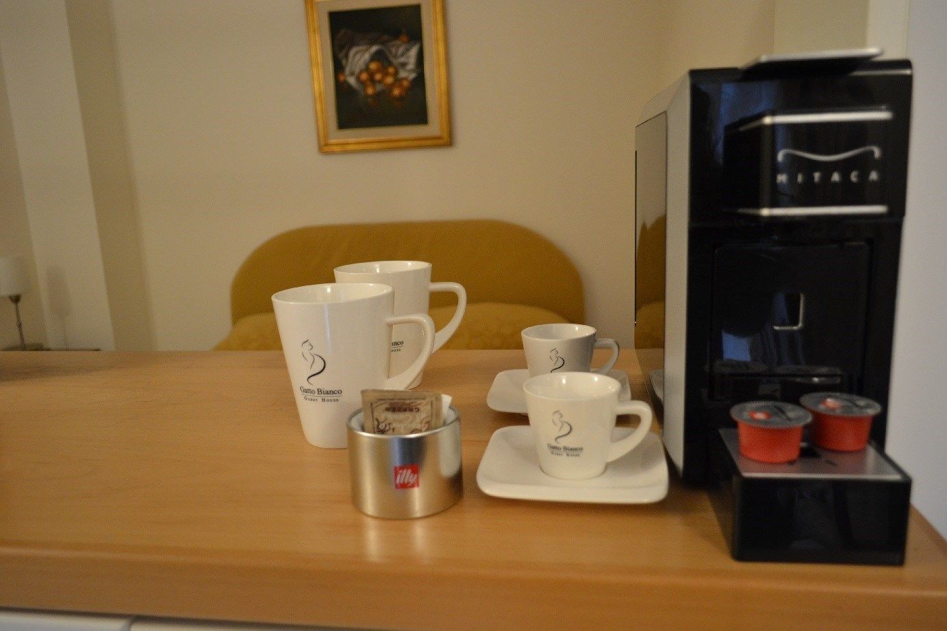 Une chambre d’hôtel équipée d’une machine à café, de deux tasses blanches ornées de logos, d’un récipient en argent et d’un tableau encadré accroché au mur.