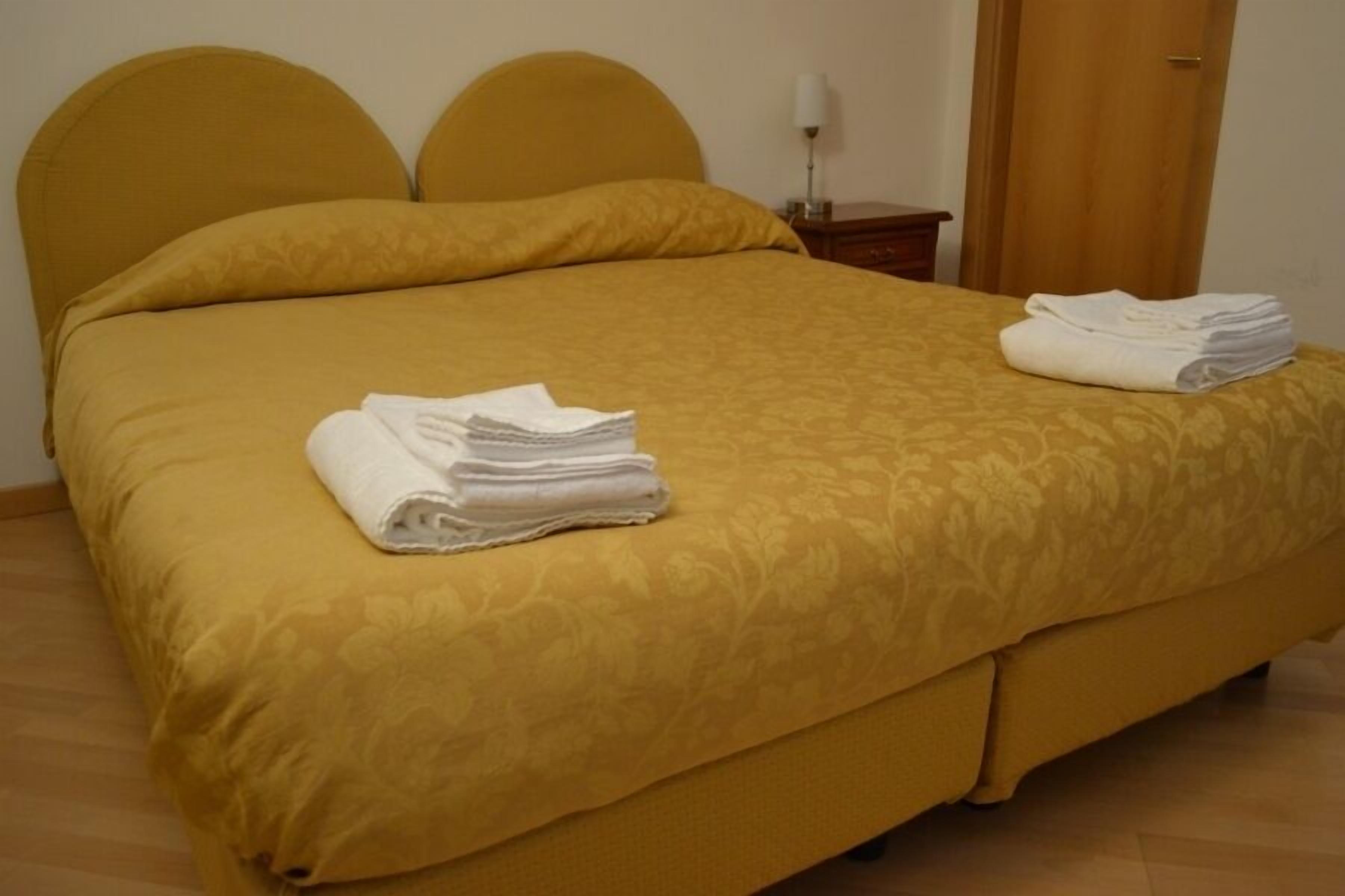 Un lit avec une tête de lit et un pied de lit jaunes, une serviette blanche pliée posée sur le lit, et une autre serviette blanche, également pliée, sur le sol, à côté du lit.