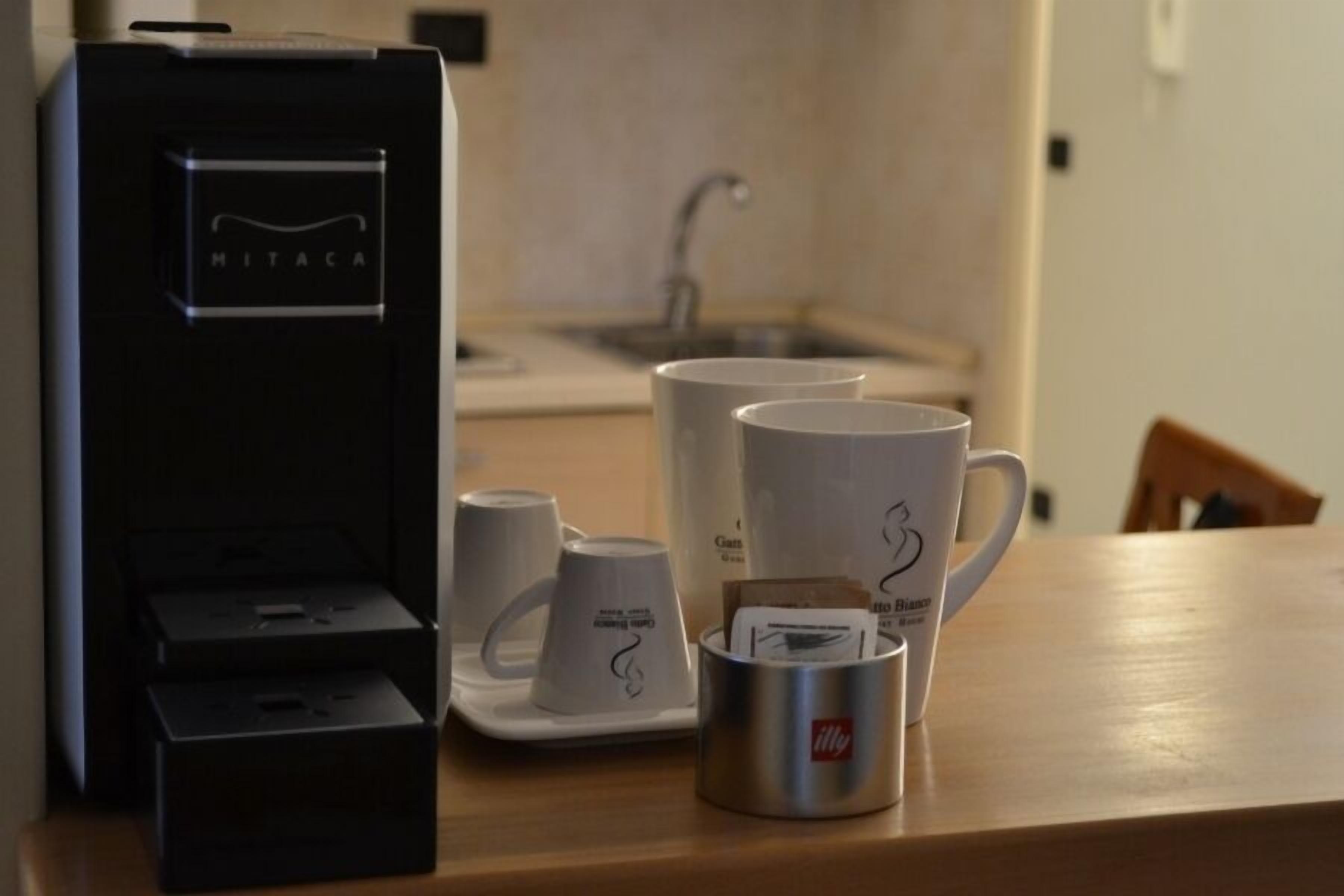 Una cafetera, tazas blancas y un vaso metálico sobre una mesa de madera en una cocina.