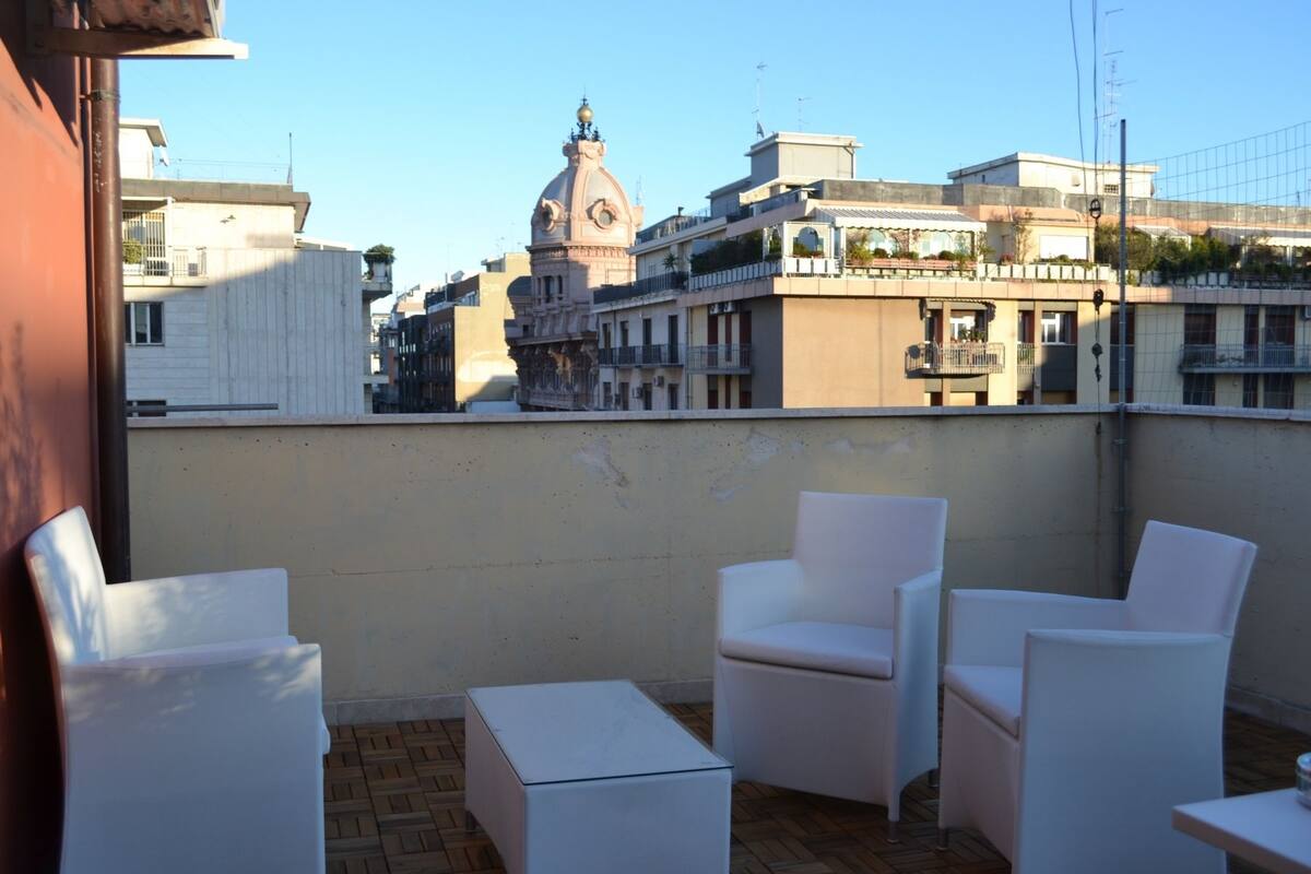 Terraço no telhado com cadeiras e mesa brancas, com vista para um horizonte urbano com edifícios históricos e uma estrutura em cúpula imponente.