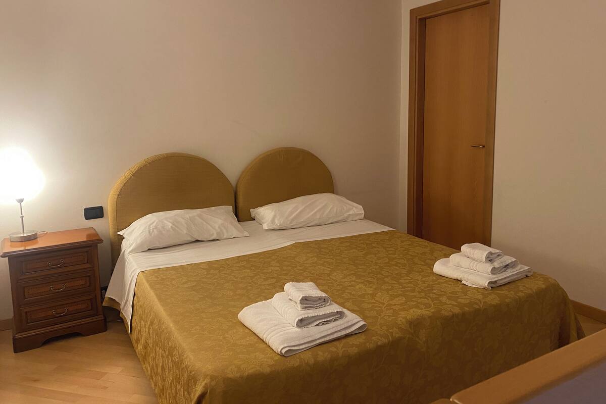 Uma cama bem arrumada com cabeceira, mesa de cabeceira com abajur e porta de madeira.