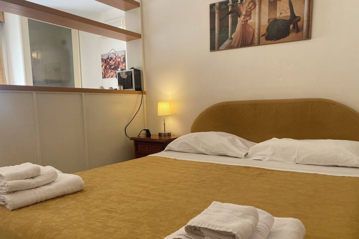 Quarto de hotel com cama, almofadas, abajur, mesa de cabeceira e um quadro na parede.