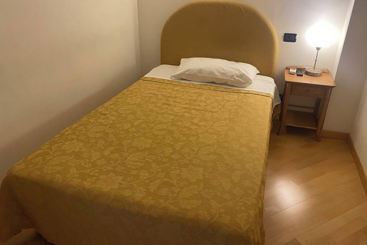 Quarto pequeno com uma cama de solteiro, cabeceira amarela, mesa de cabeceira com abajur e telefone.