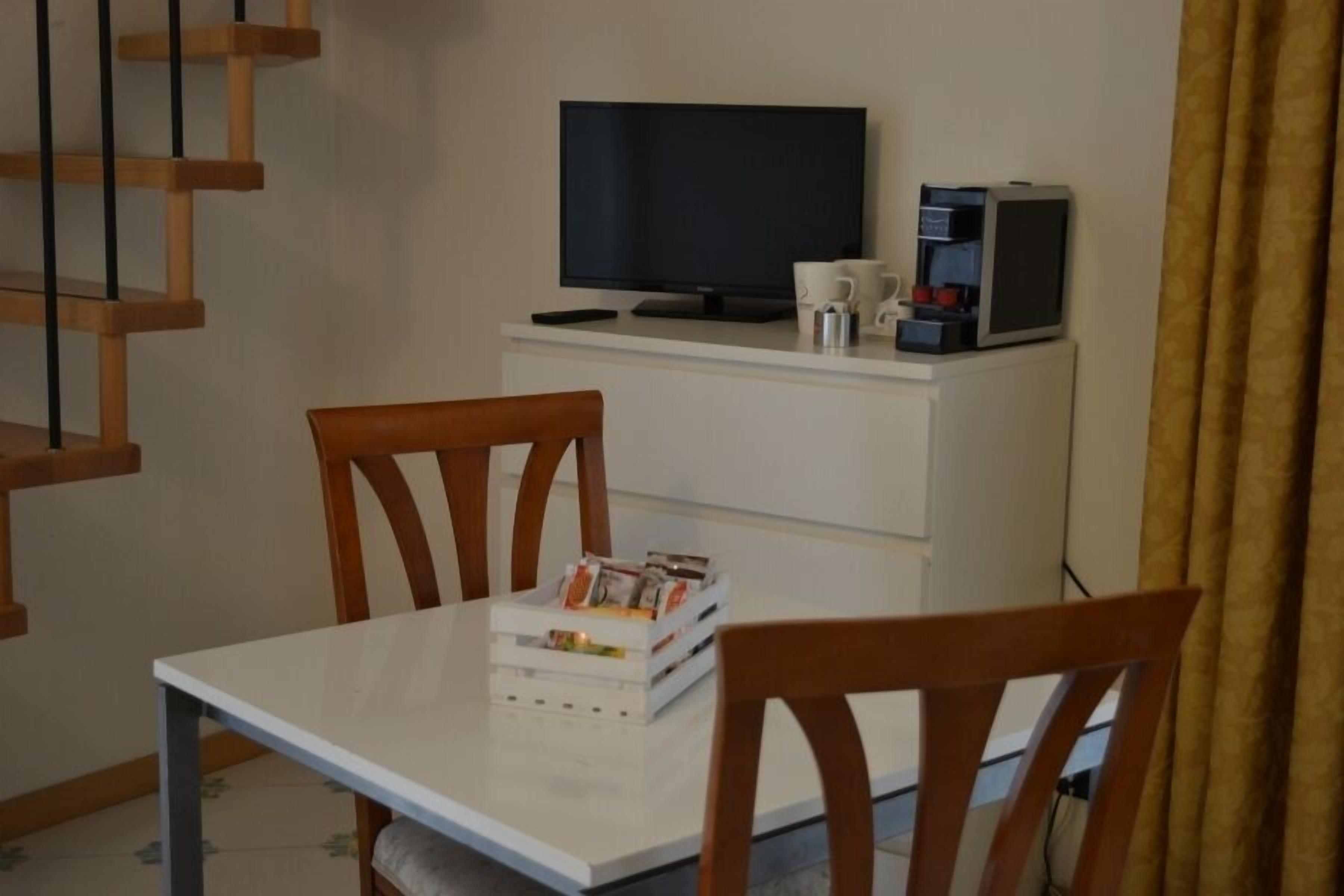 Ein kleiner Raum mit einem weißen Tisch, zwei Holzstühlen, einem Fernseher, einer Kaffeemaschine und einem kleinen Schrank.