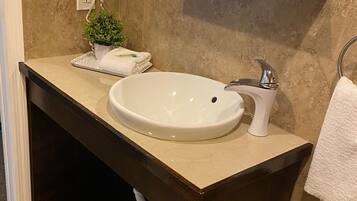 Een badkamer met een beige wastafel, een haardroger, een kleine potplant en een prullenbak.