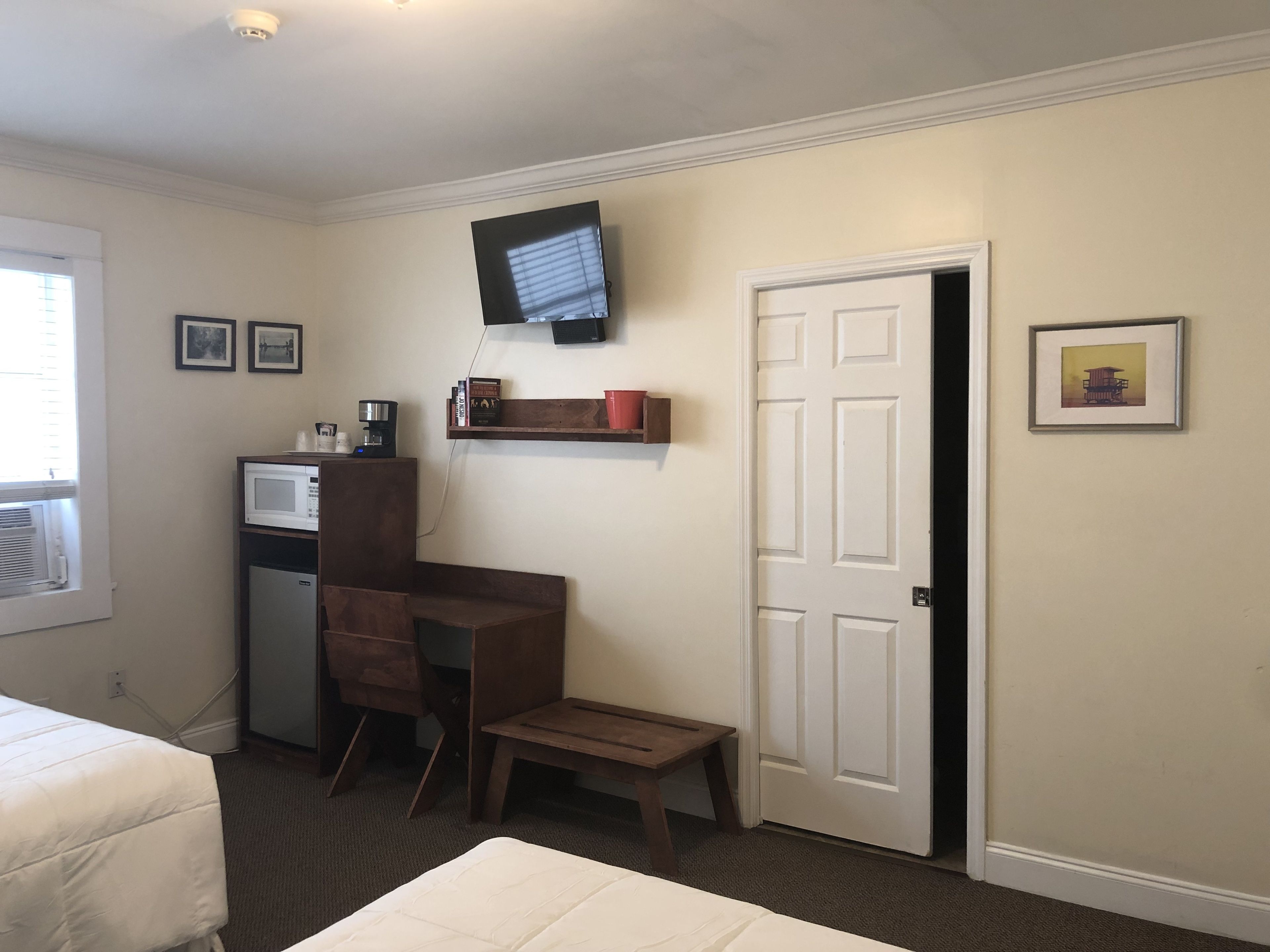 A hotel room with a bed, a desk with a chair, a television mounted on the wall, a door, and a small shelf with books and a red cup.