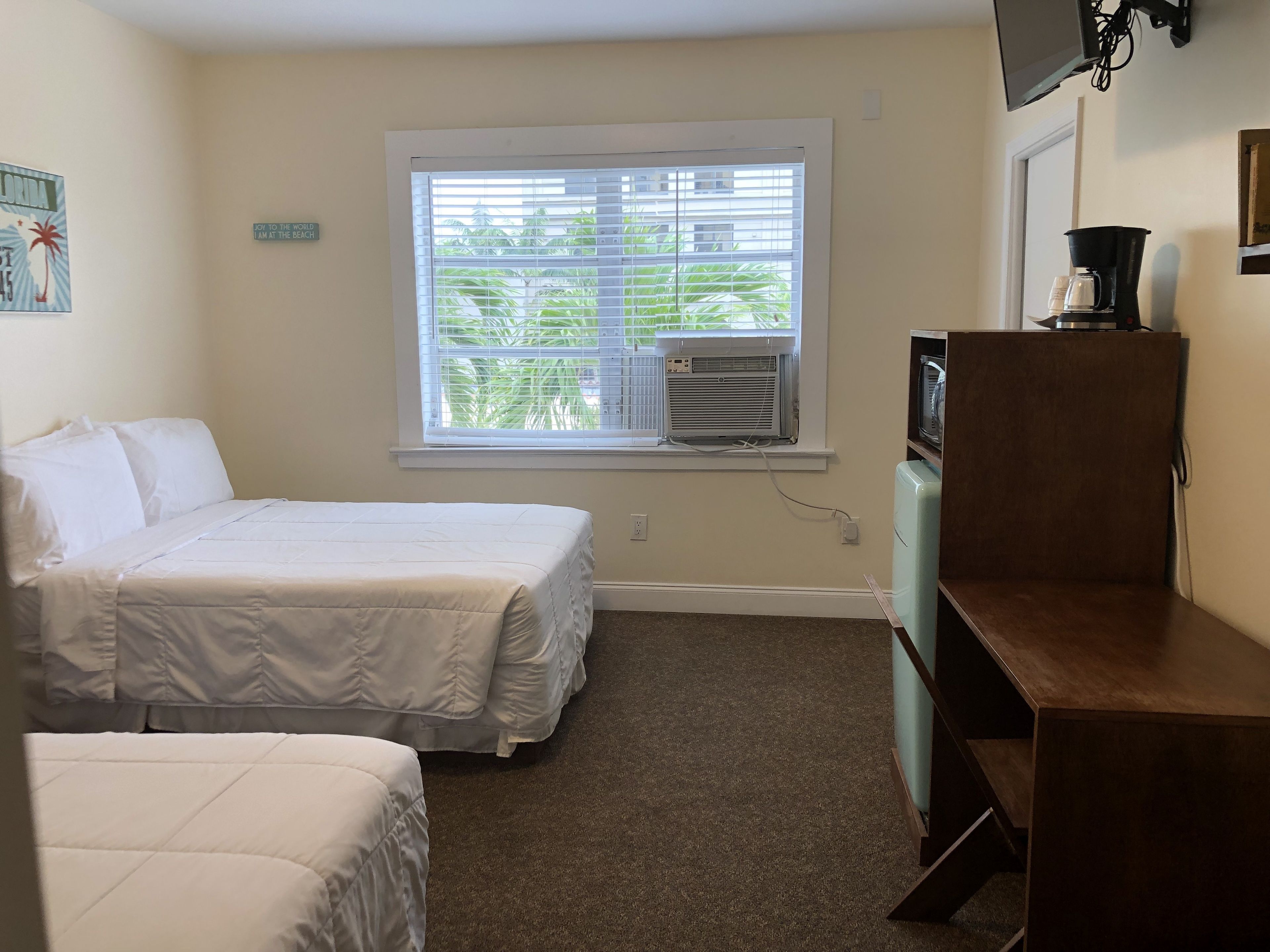 A hotel room with two beds, a window with blinds, an air conditioning unit, a coffee maker, and a small wooden desk.