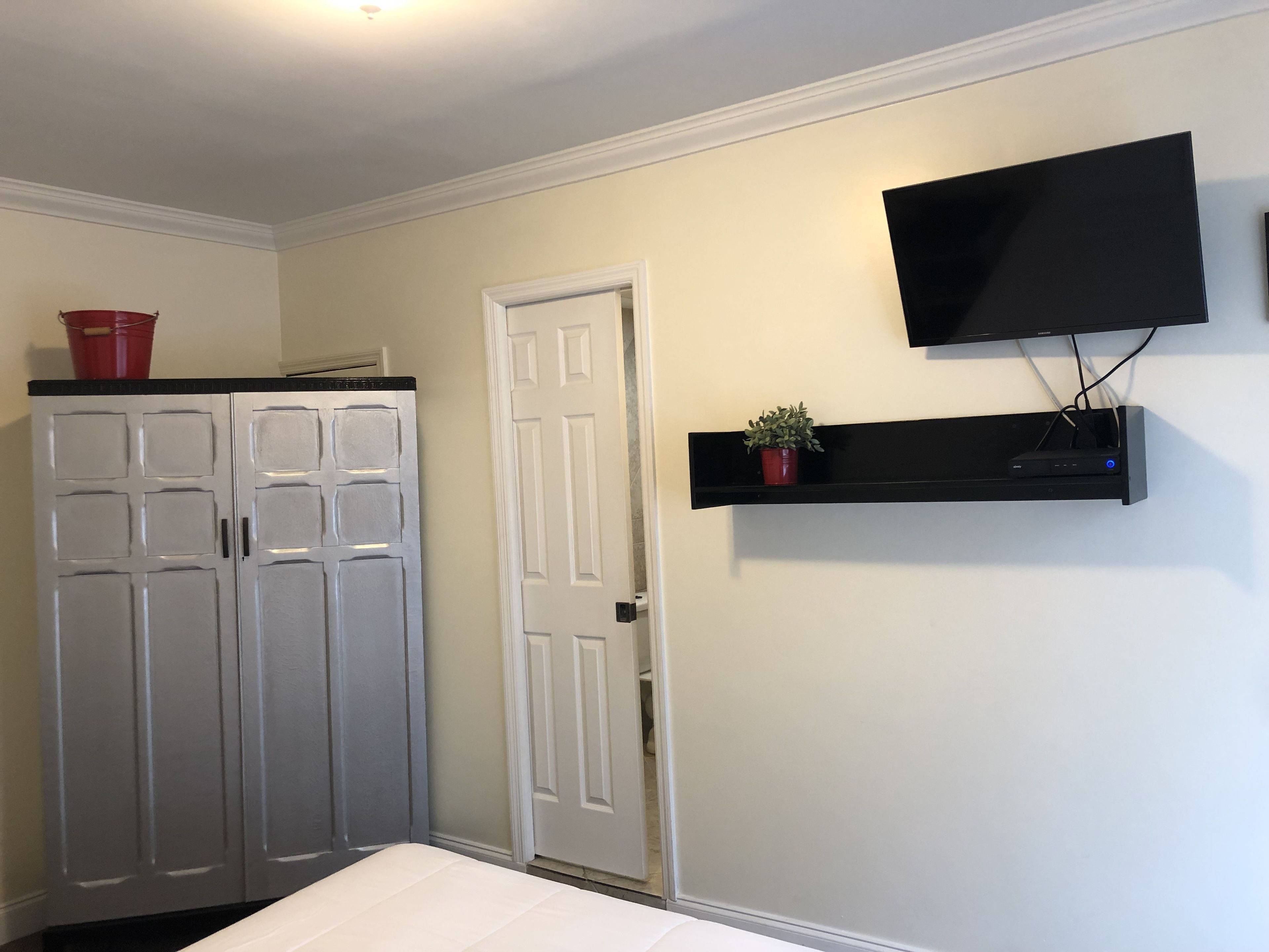 Une chambre à coucher comprenant un lit, une armoire, une porte, un téléviseur fixé au mur et une étagère avec une plante.